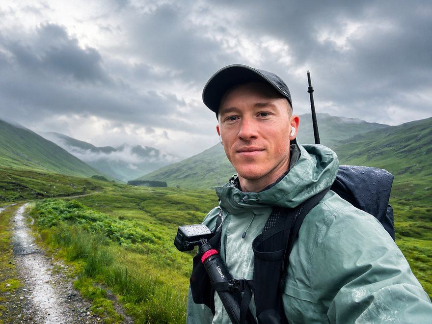 Jack Brookes on a wet gravel track through a Scottish glen, pack and chest-mounted camera on, low cloud over the hills
